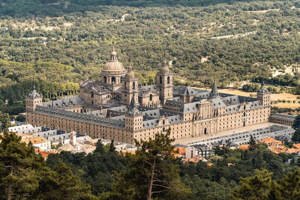 Vista aérea del monasterio de El Escorial, con su cúpula y torres, rodeado de bosque y casas del pueblo.