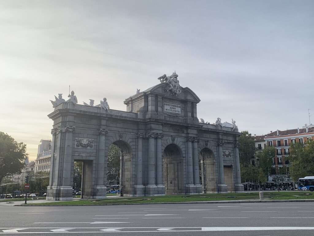 Puerta de Alcalá de Madrid al atardecer, con esculturas en lo alto, una rotonda con césped y un autobús al fondo.