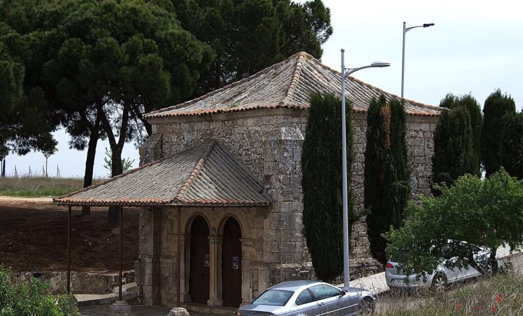 Ermita de piedra con porche de teja, rodeada de pinos y cipreses, con farolas y coches aparcados junto a la calle
