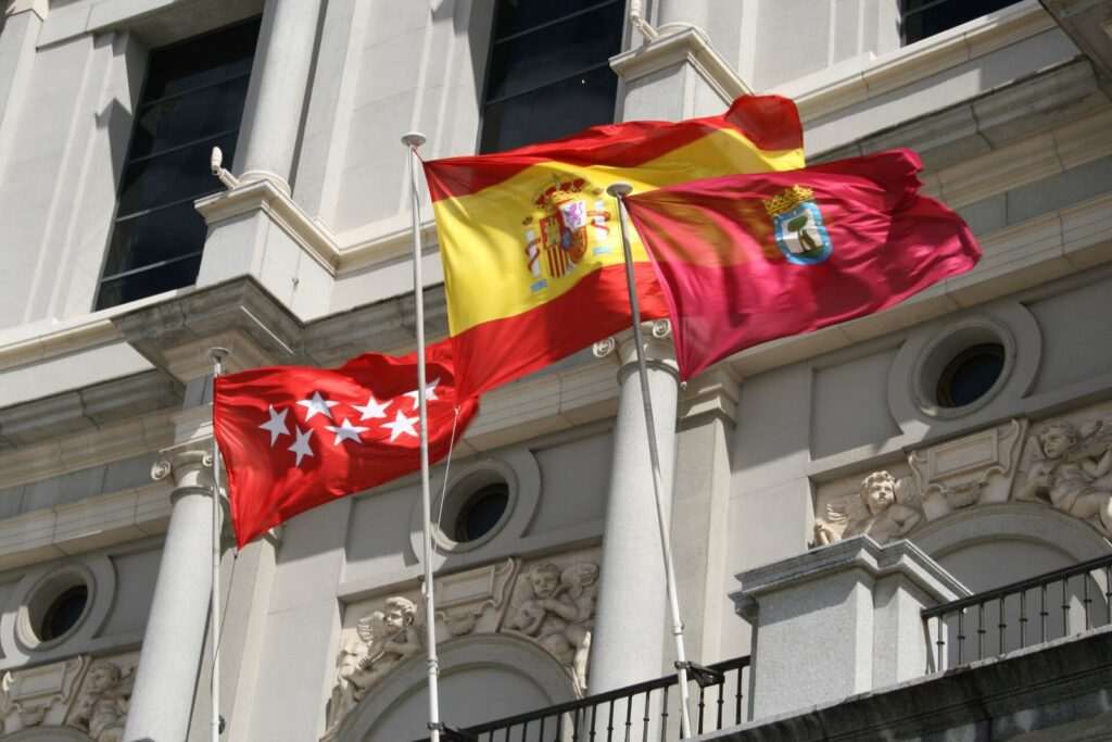 Banderas de la Comunidad de Madrid, de España y de la ciudad de Madrid ondeando ante la fachada de un edificio de piedra